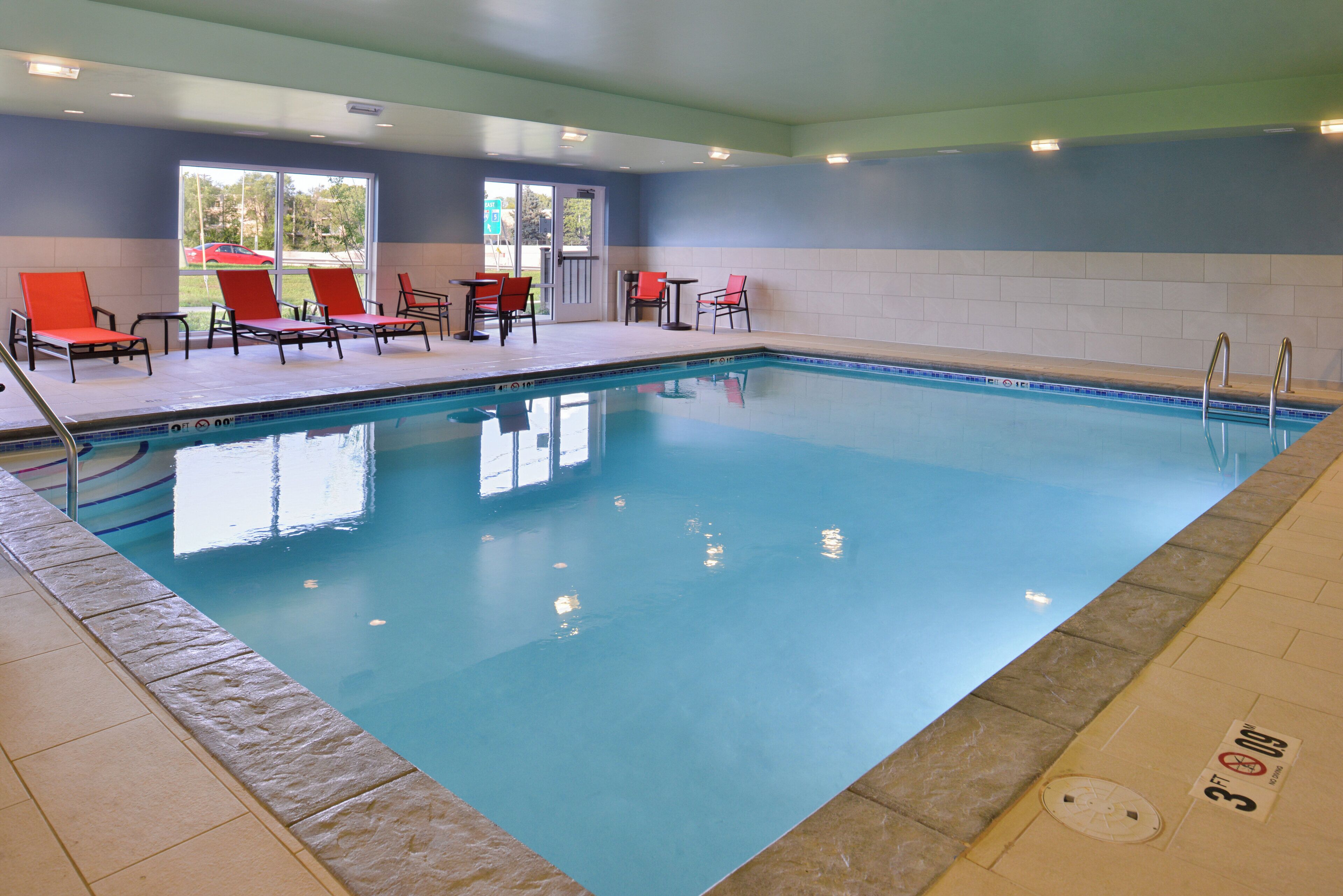An indoor swimming pool with clear blue water, surrounded by red and black lounge chairs and tables.