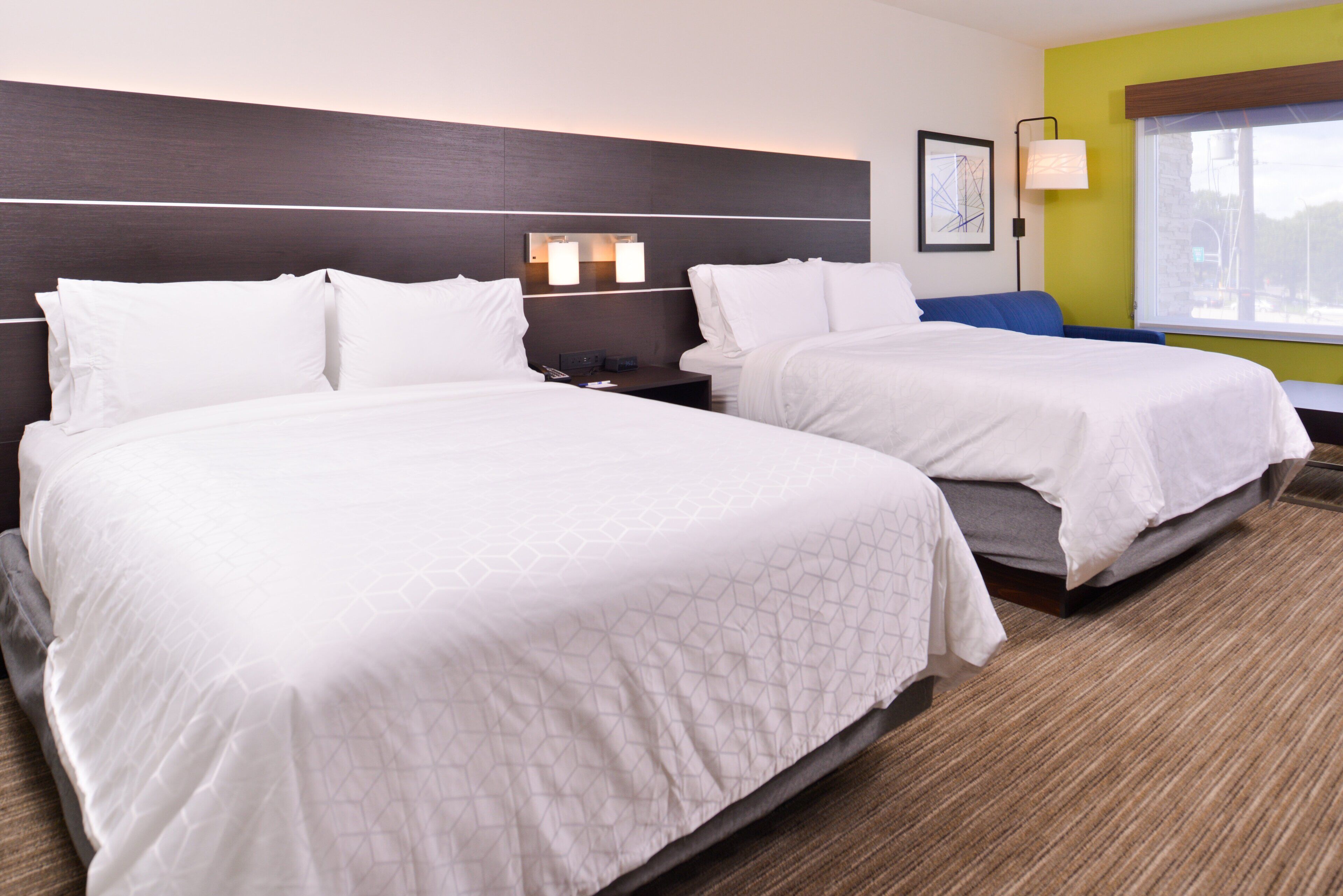 A hotel room with two beds, a nightstand, a lamp, and a window with a view.