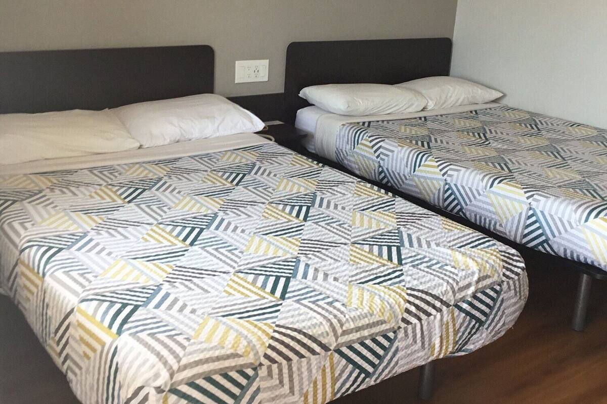 Quarto de hotel com duas camas, cada uma com roupa de cama estampada e cabeceira. Há uma luminária fixada na parede acima das camas e um detector de fumaça na parede.