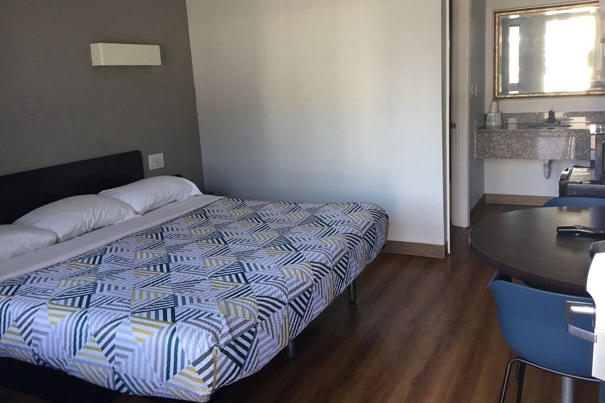 Quarto de hotel com cama, uma pequena mesa e área de cozinha compacta.