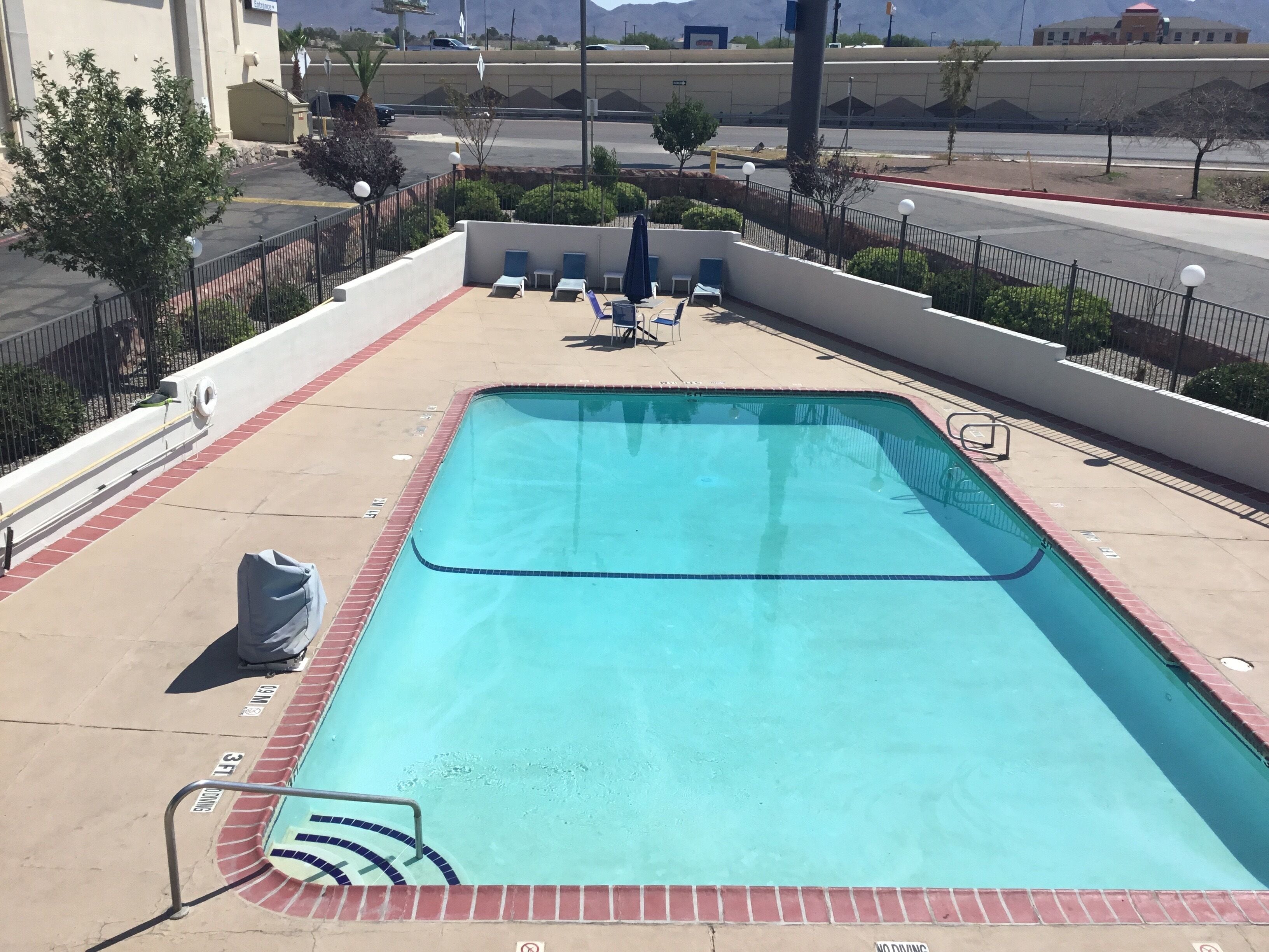 Una piscina rectangular con borde de azulejos, rodeada por un muro de ladrillo bajo y una barandilla metálica. Hay sillas de descanso y una mesita pequeña al borde de la piscina.
