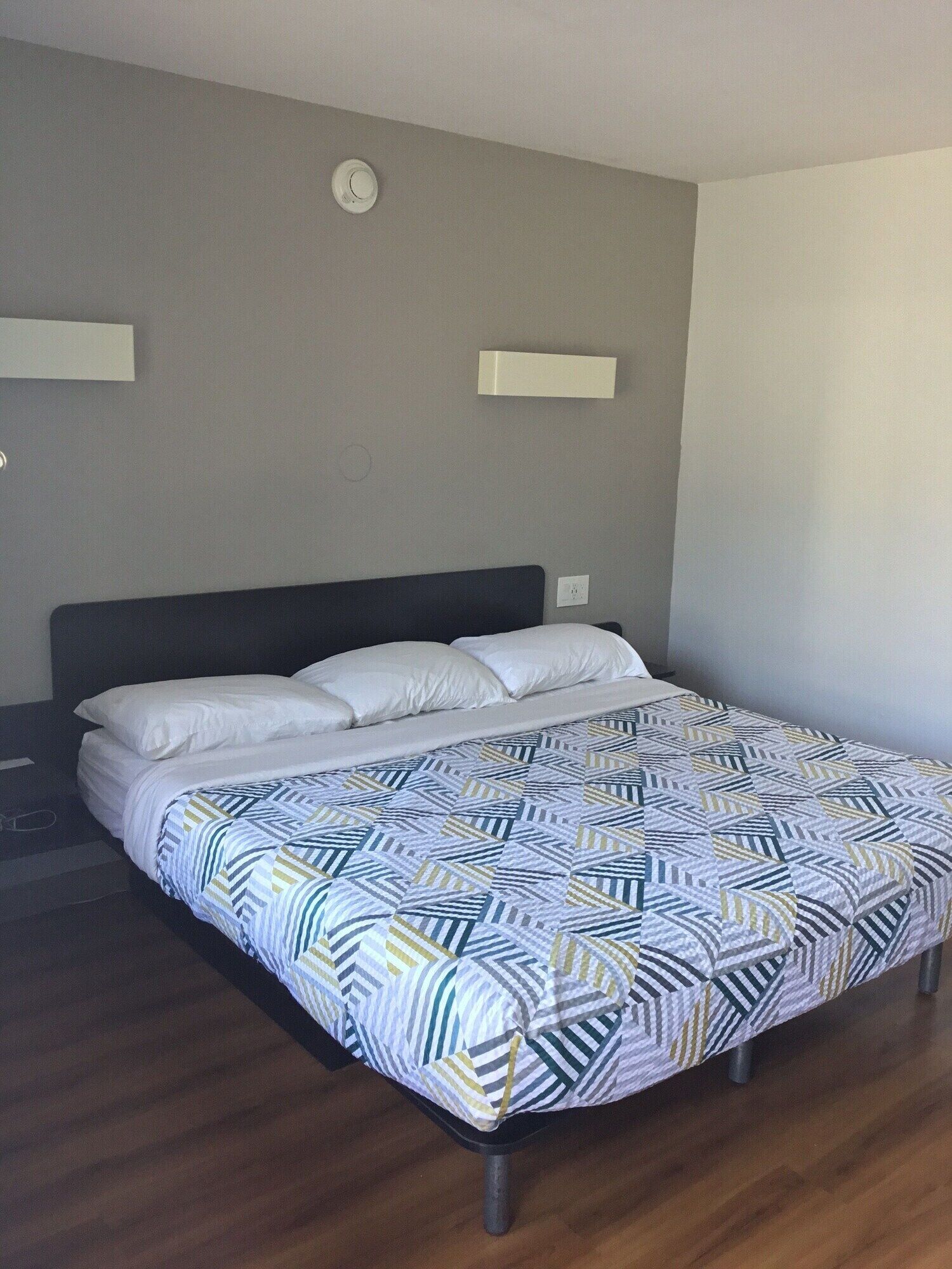 Una cama bien tendida con una colcha estampada, un cabecero oscuro y una pared lisa con un interruptor de luz y un detector de humo.