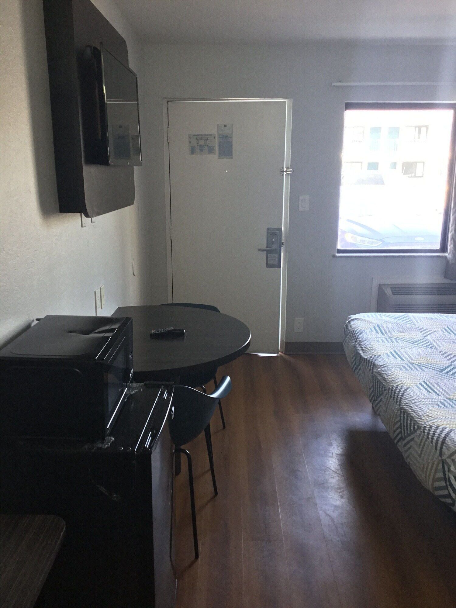 Een kleine kamer met een bed, een eettafel met stoelen, een magnetron, een aan de muur bevestigde televisie en een deur met een raam.