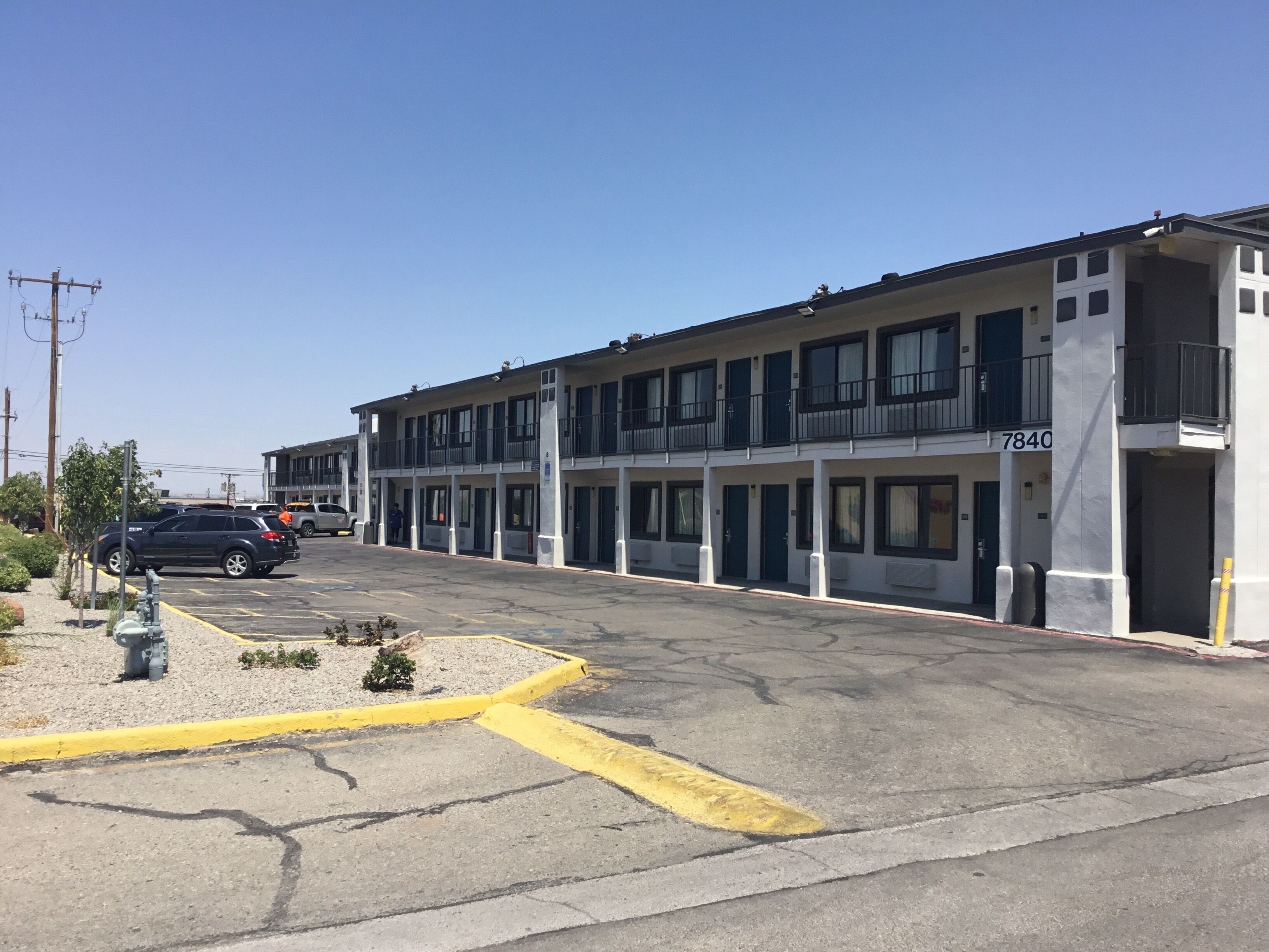 A multi-story motel with a row of rooms, a parking lot, and a clear blue sky.