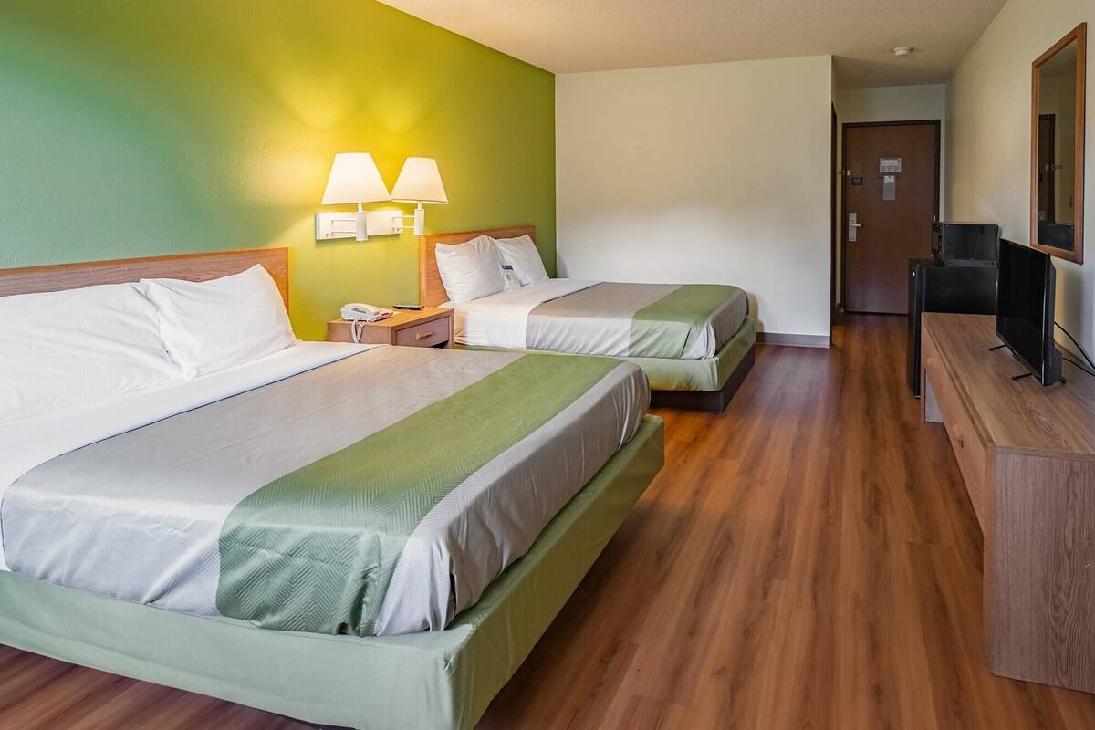 A hotel room with two beds, a wooden floor, a desk with a television, and a green wall with two wall lamps.