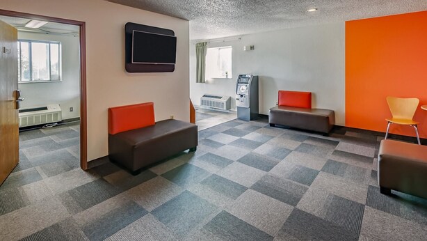 A modern waiting area with two cushioned seating areas, a flat-screen TV mounted on the wall, and a small ATM machine. - Motel 6 Drums, PA (Drums)