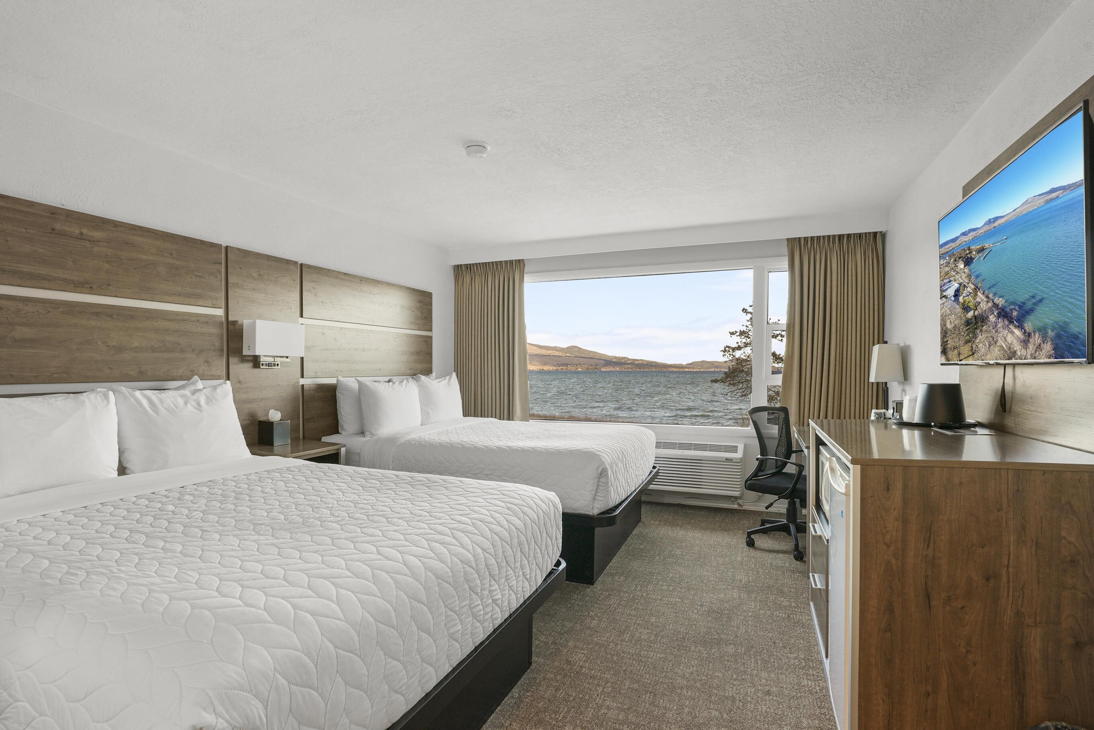 Habitación de hotel con dos camas, un escritorio, una silla y un amplio ventanal con vista al agua.