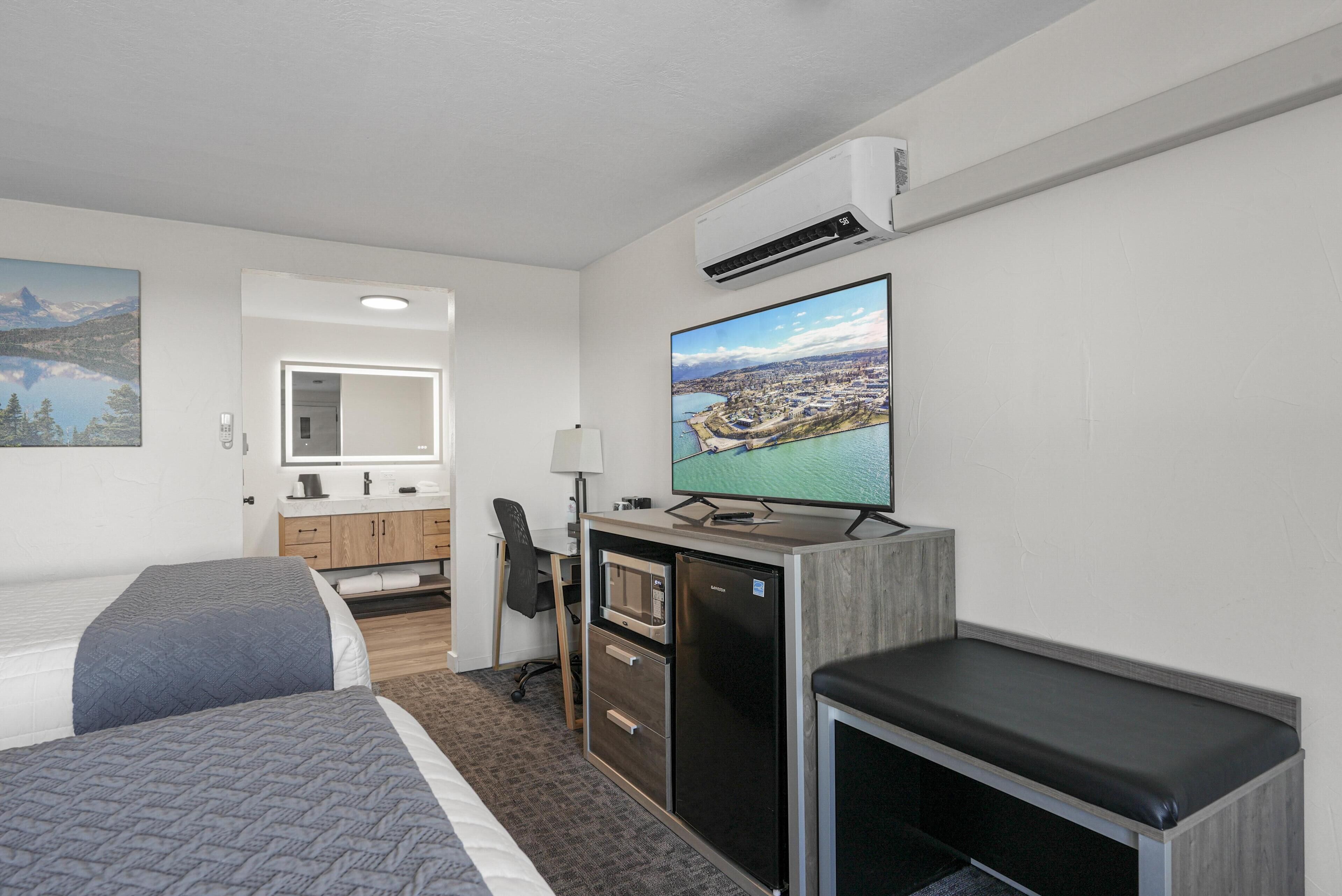 A hotel room with two beds, a desk with a computer, a refrigerator, a microwave, a bench, and a large TV.