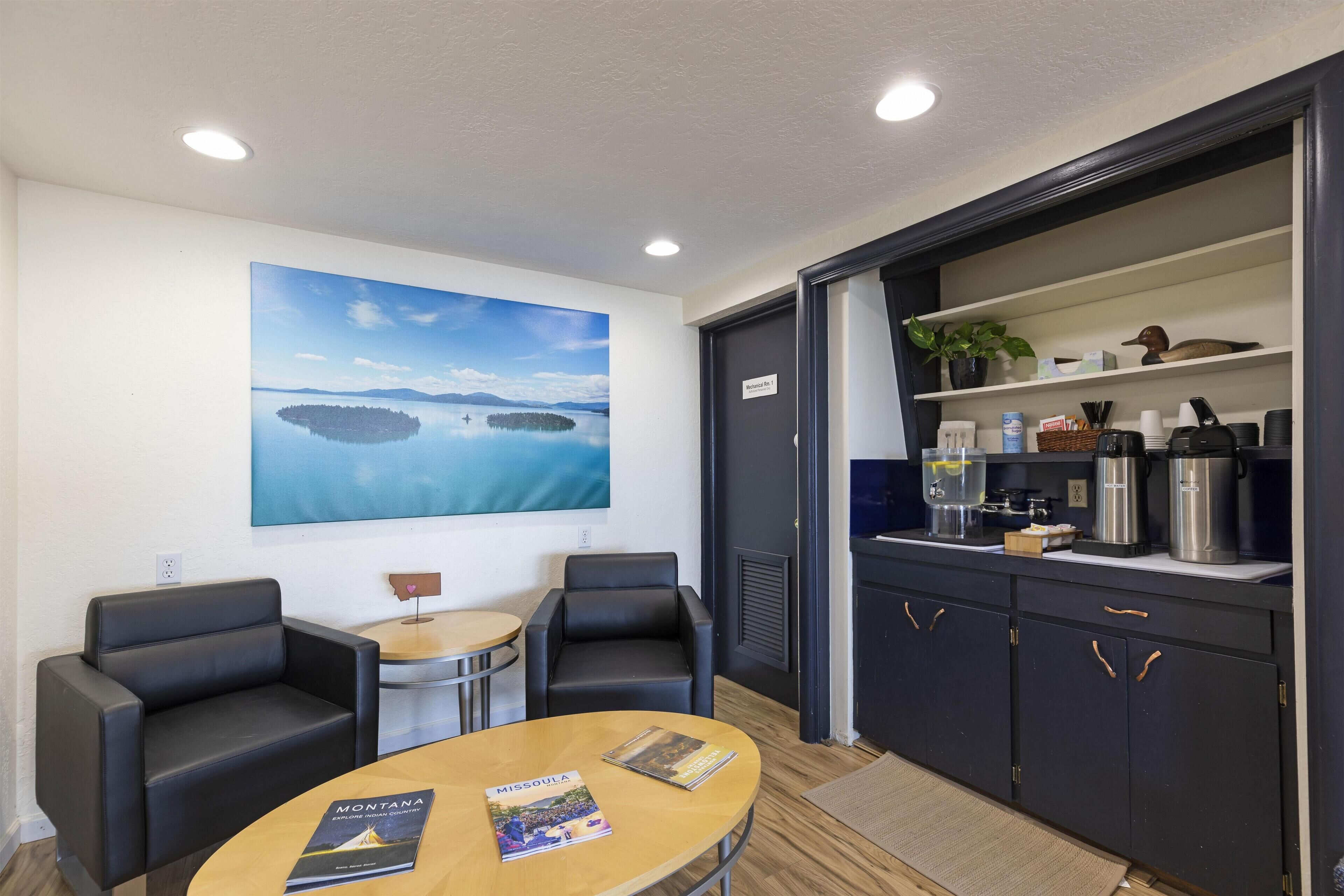 Una habitación moderna con una mesa redonda, dos sillones de cuero negro, una estantería con diversos objetos y un cuadro de un lago en la pared.