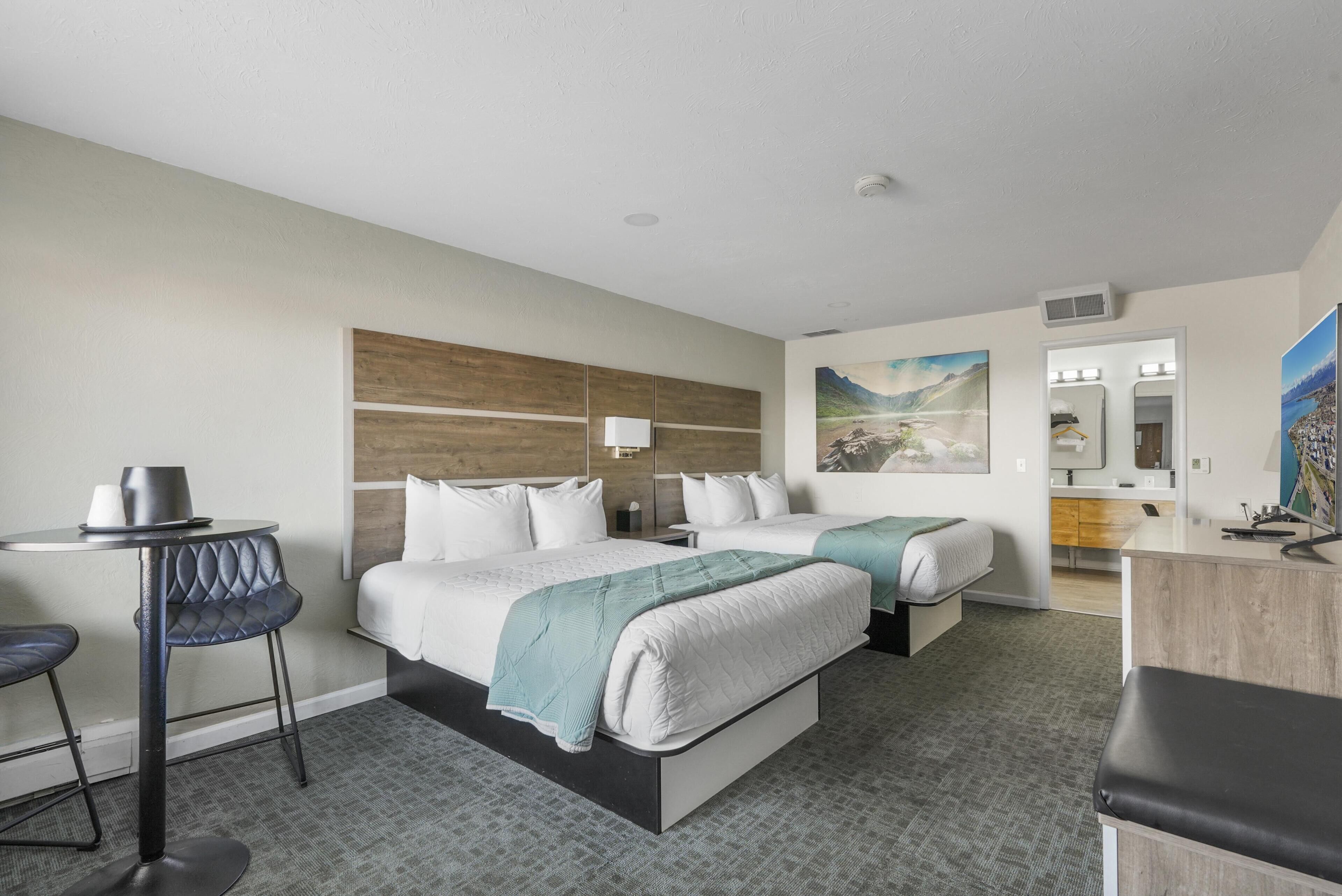 A hotel room with two beds, a desk, a chair, a small table, and a bathroom visible through an open door.