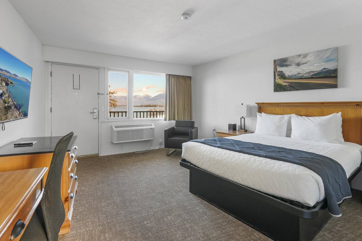 A hotel room with a bed, a desk, a chair, a window with a view, and a painting on the wall.