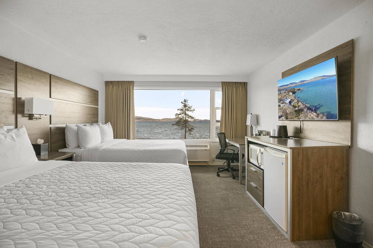 A hotel room with two beds, a desk, a chair, a small kitchenette, and a large window with a view of the sea and hills.