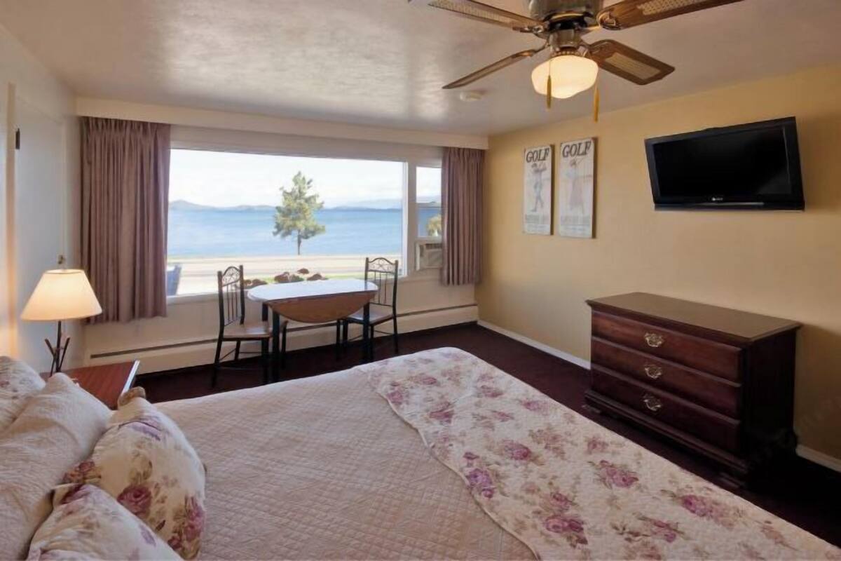 A hotel room with a bed, a desk, a chair, a dresser, a television, and a view of the sea.