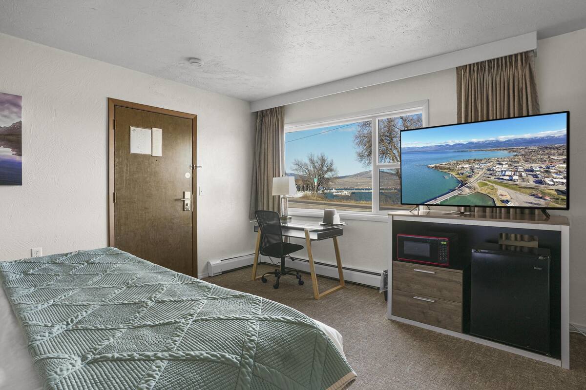 A hotel room with a bed, desk, chair, TV, and a view of a coastal town.