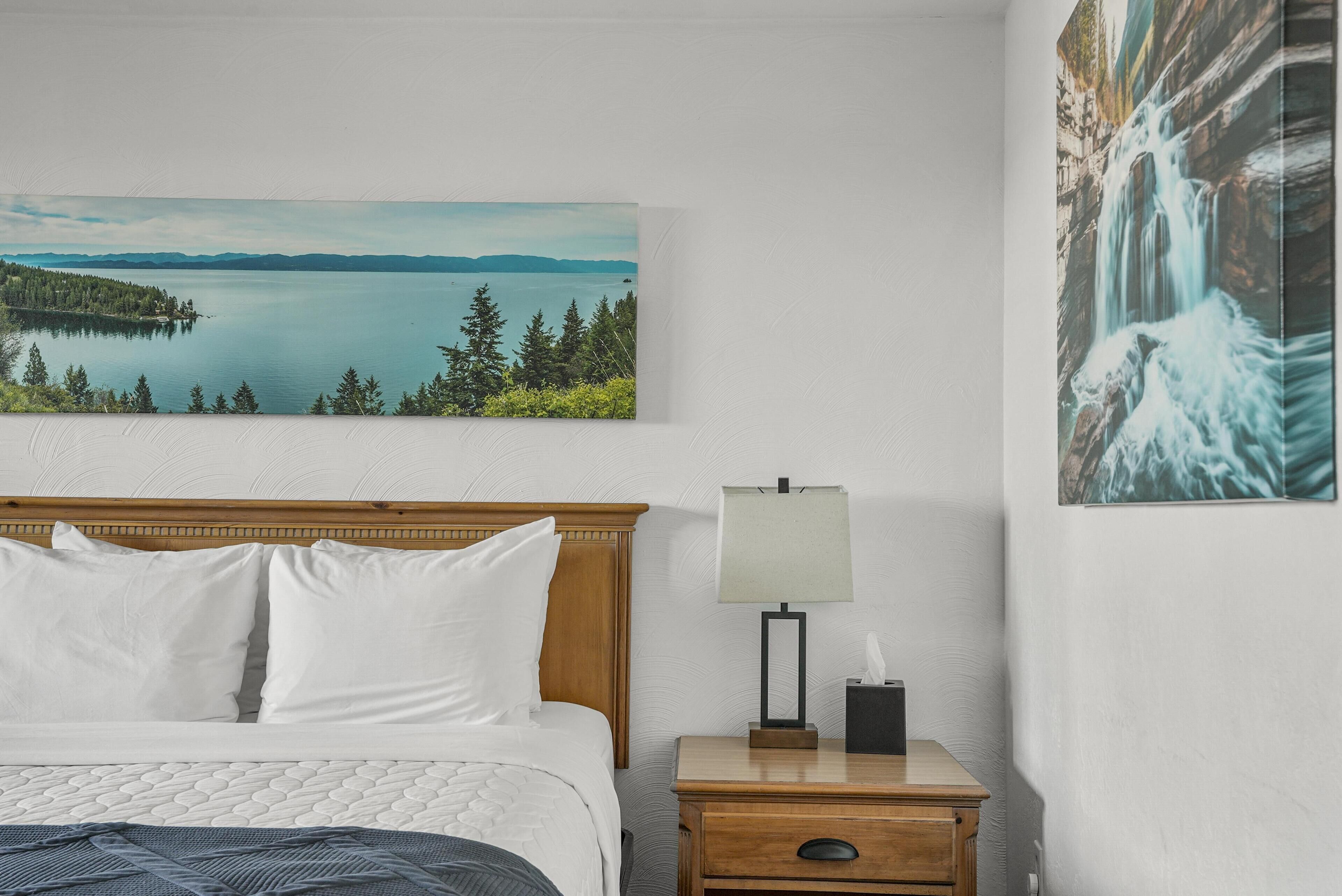 A hotel room with a bed, a nightstand with a lamp, and two large landscape paintings on the walls.