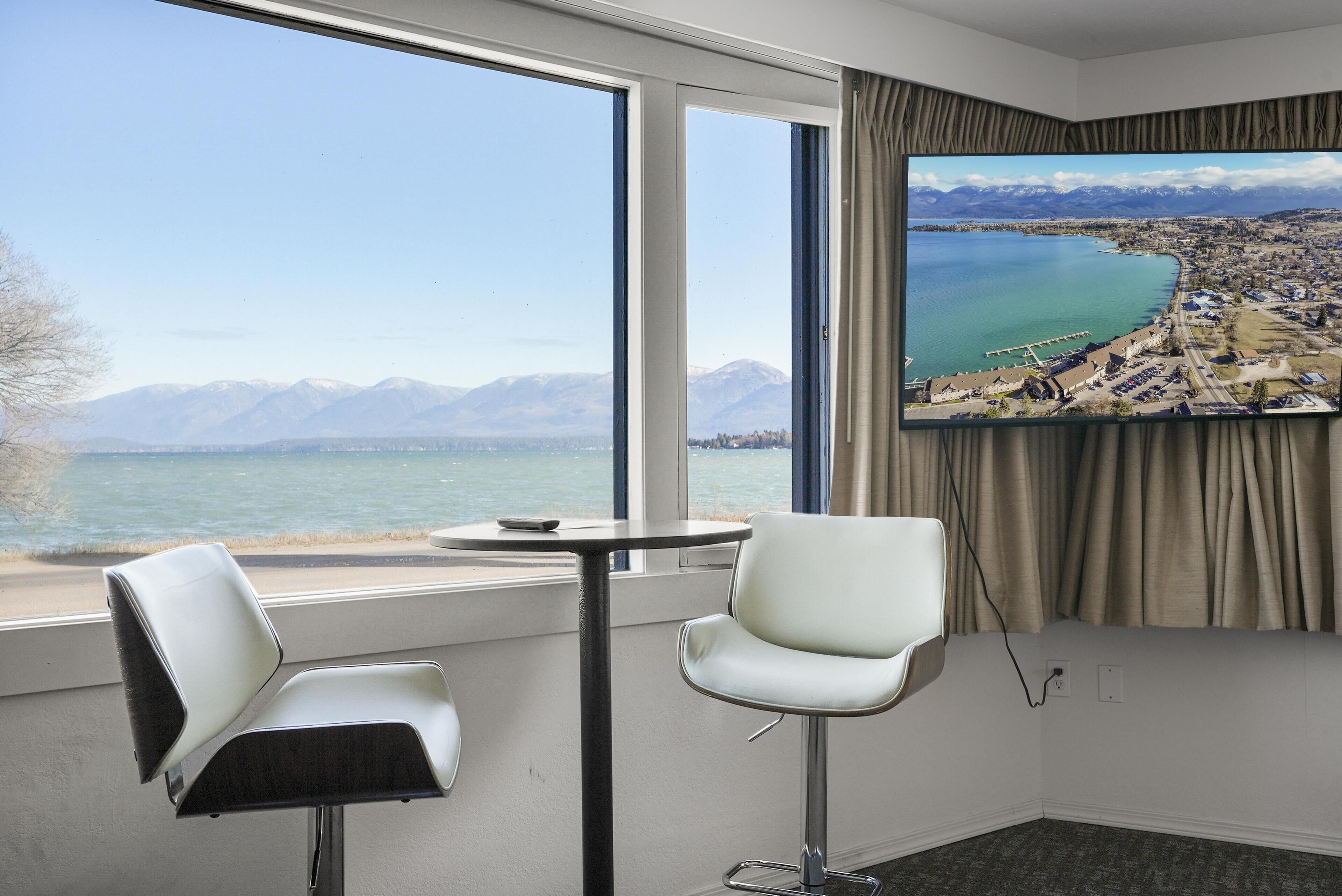 Ein Hotelzimmer mit großem Fenster und einem schönen Blick auf einen See und Berge. Zwei weiße Stühle und ein kleiner runder Tisch sind vorhanden.