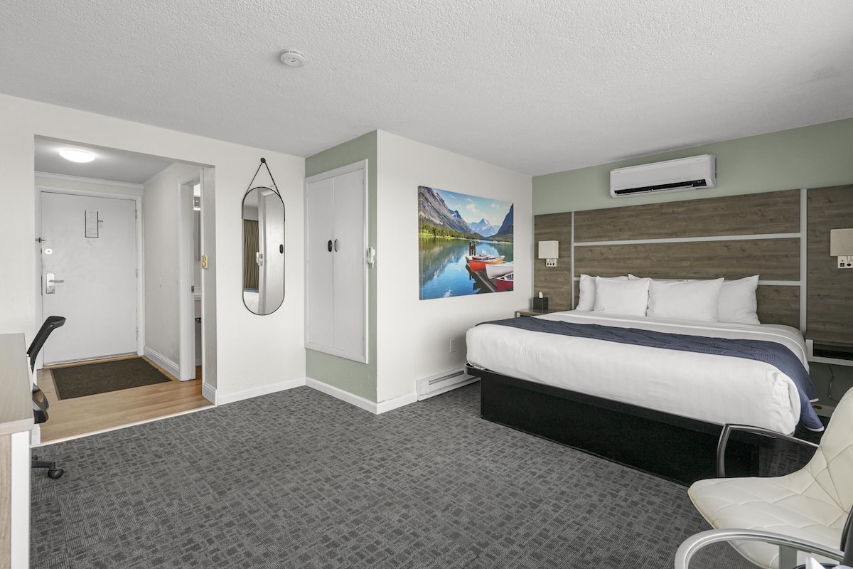 A hotel room with a bed, a desk, a chair, a painting, and an air conditioning unit.