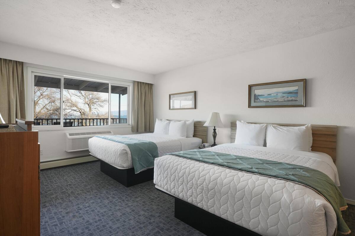 A hotel room with two beds, a large window with a view, a desk, and a lamp.