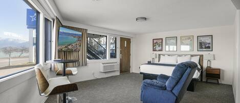 A hotel room with a bed, a blue reclining chair, a small table, and a window with a view of a coastal area.