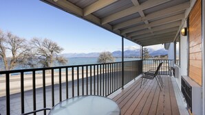 Una terraza de madera con una mesa y sillas de vidrio, con vistas a un cuerpo de agua y montañas.