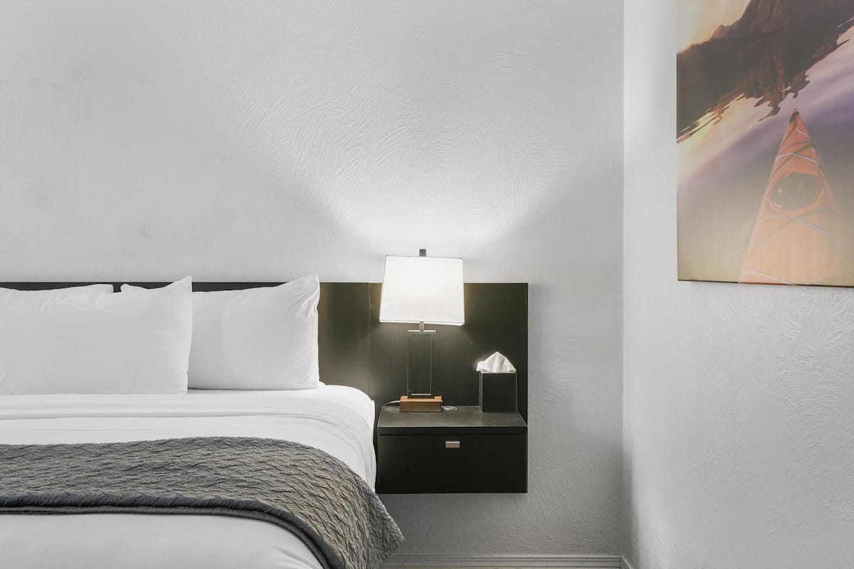 A neatly made bed with white linens and a gray throw blanket. A black bedside table with a white lamp and a tissue box is positioned next to the bed.