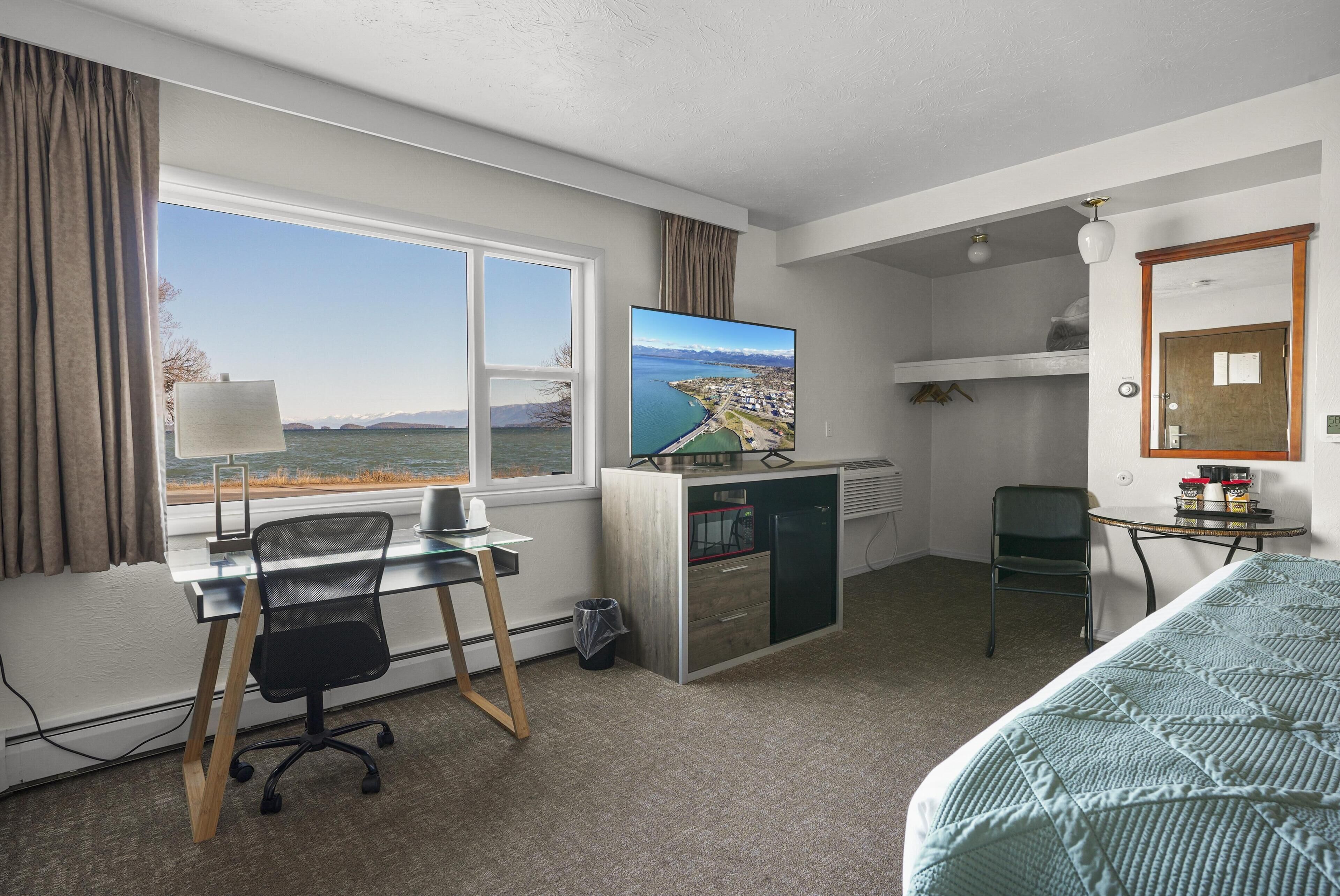 A hotel room with a desk, chair, bed, TV, and a view of a lake.