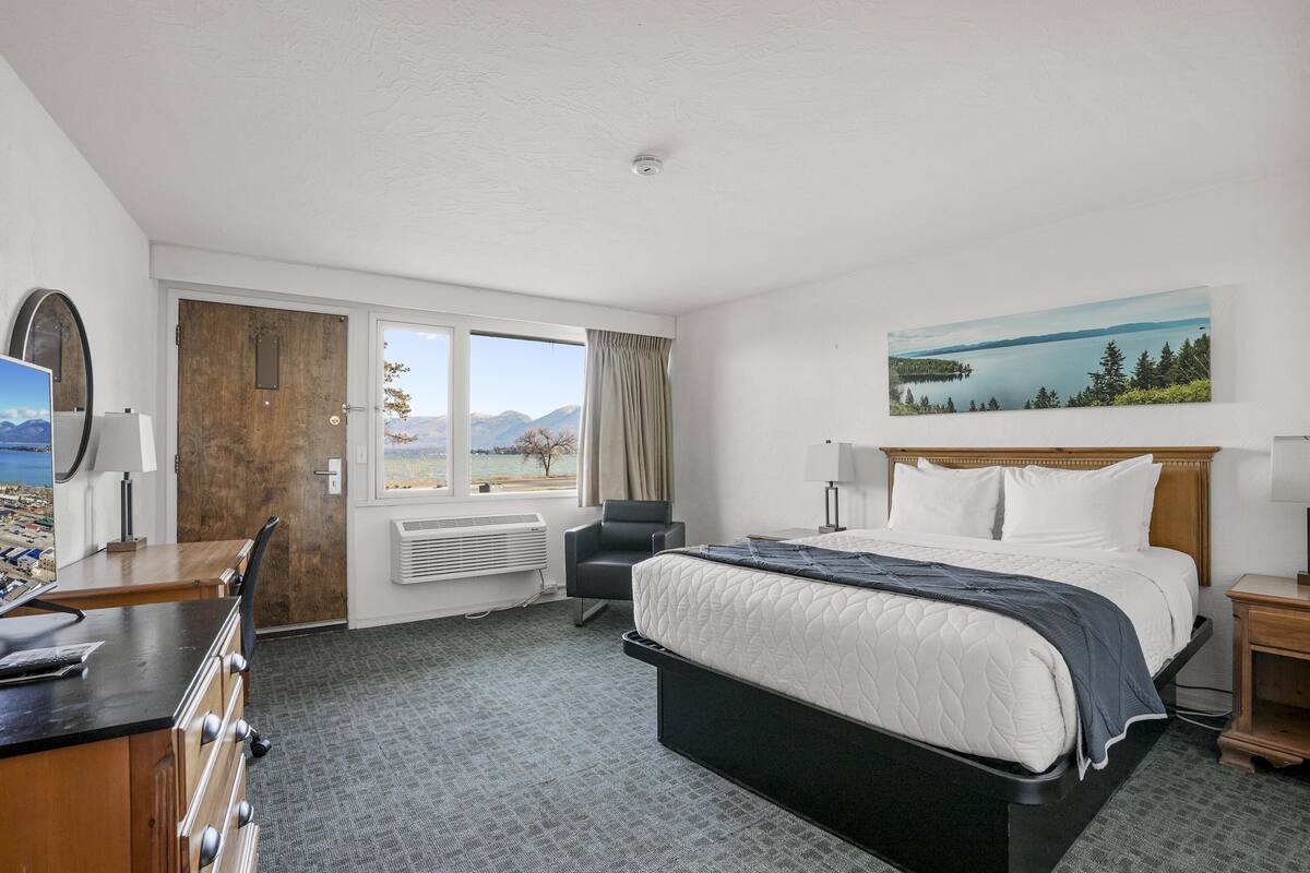 A hotel room with a bed, desk, chair, and a painting of a lake.