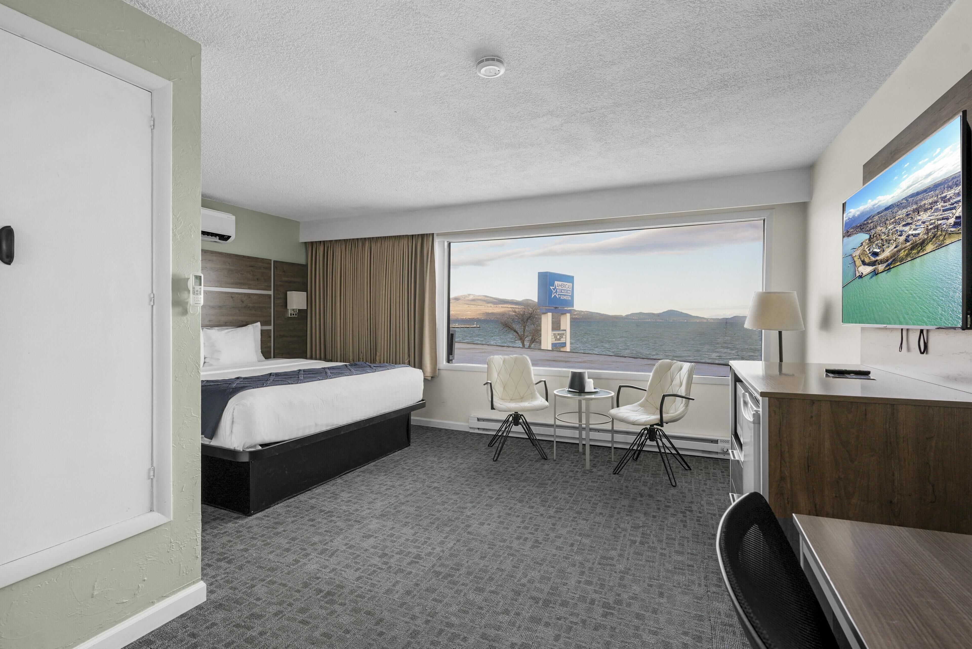 Een hotelkamer met een bed, een bureau, een stoel, een kleine tafel en een groot raam met uitzicht op een stad en bergen.
