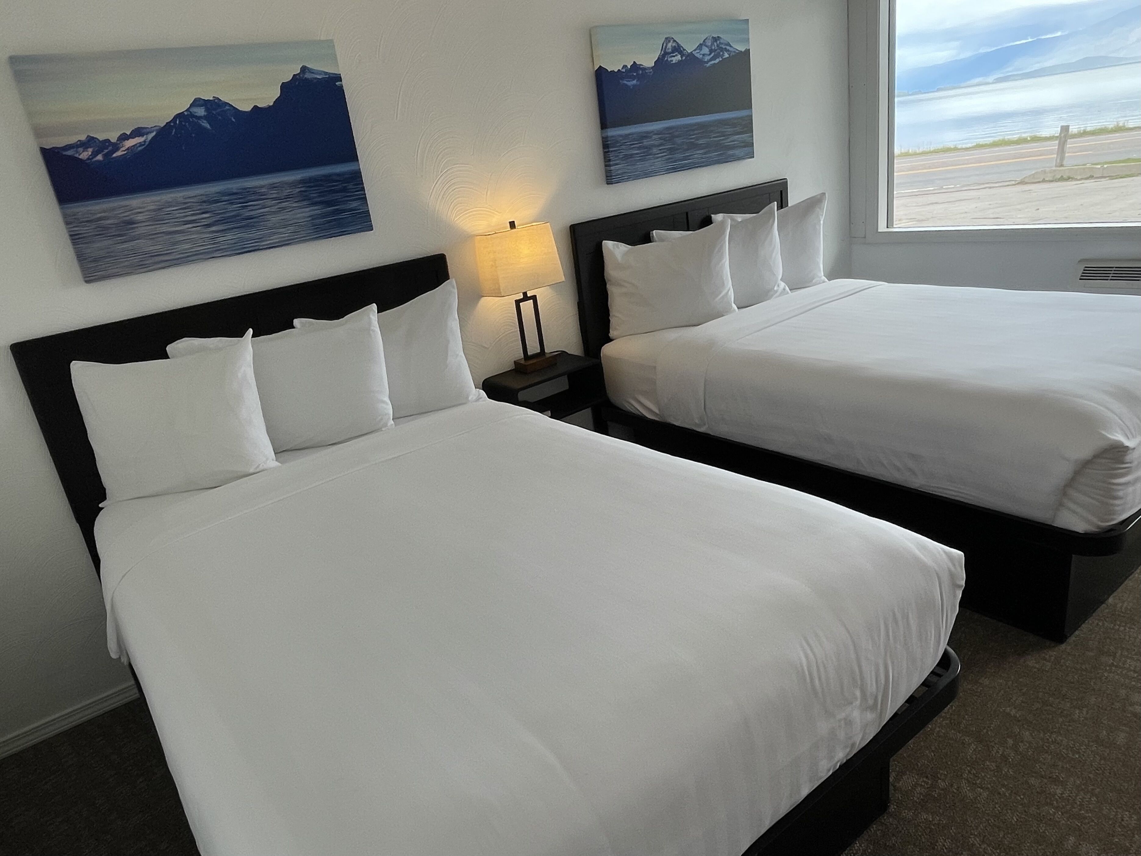Habitación de hotel con dos camas, una mesita de noche con lámpara, y dos cuadros de montañas y agua en la pared.