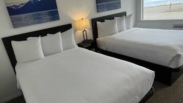 Habitación de hotel con dos camas, una mesita de noche con lámpara, y dos cuadros de montañas y agua en la pared.