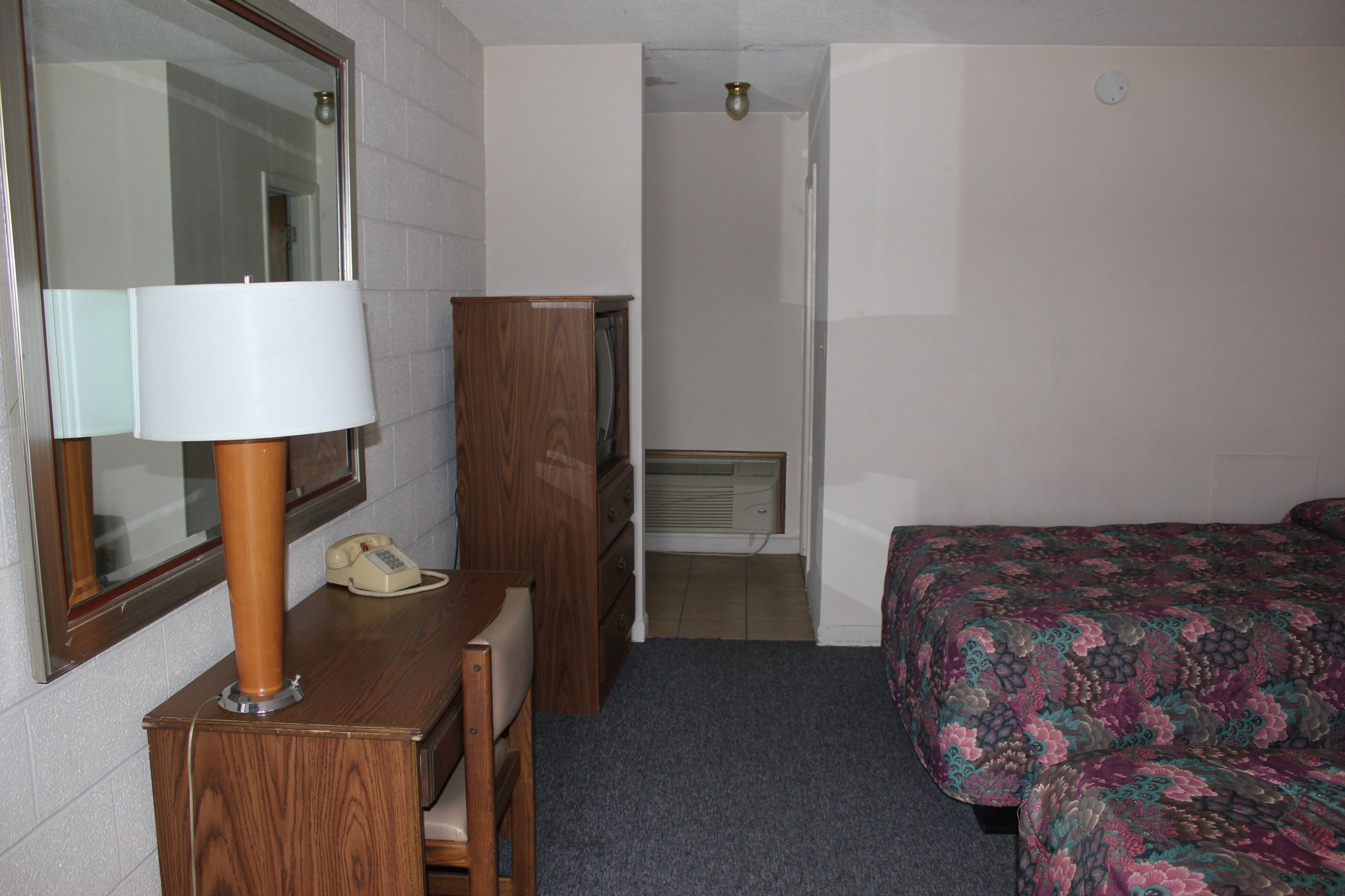 Une chambre d’hôtel équipée d’un bureau, d’un téléphone, d’un miroir, d’une armoire, d’un lit avec une couvre-lit à motifs floraux et d’une porte donnant sur une salle de bain.