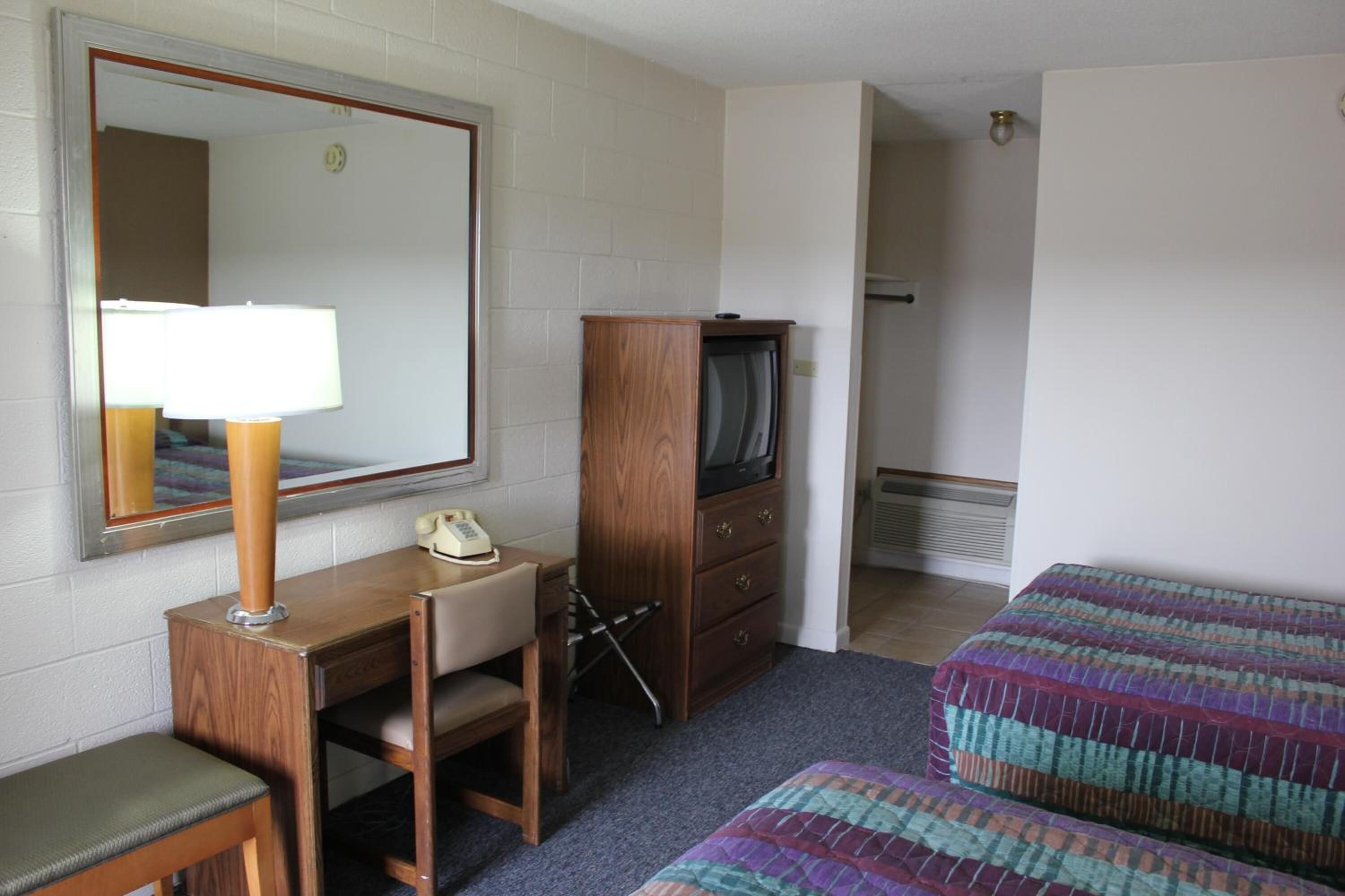 A compact hotel room with a wooden desk, a chair, a television, a bed with a colorful blanket, and a mirror on the wall.