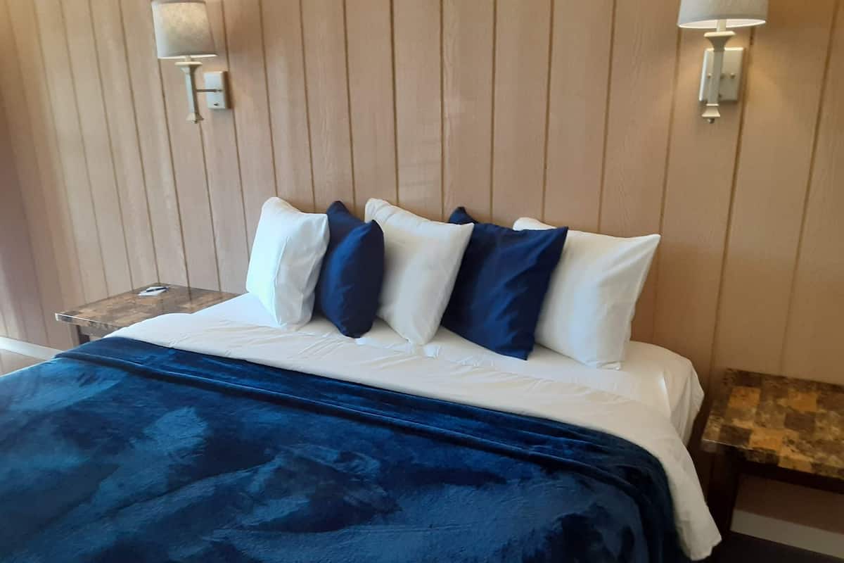 A bed with a white and blue duvet, a dark blue blanket, and several pillows in white and blue. There are two wall-mounted lamps on either side of the bed.