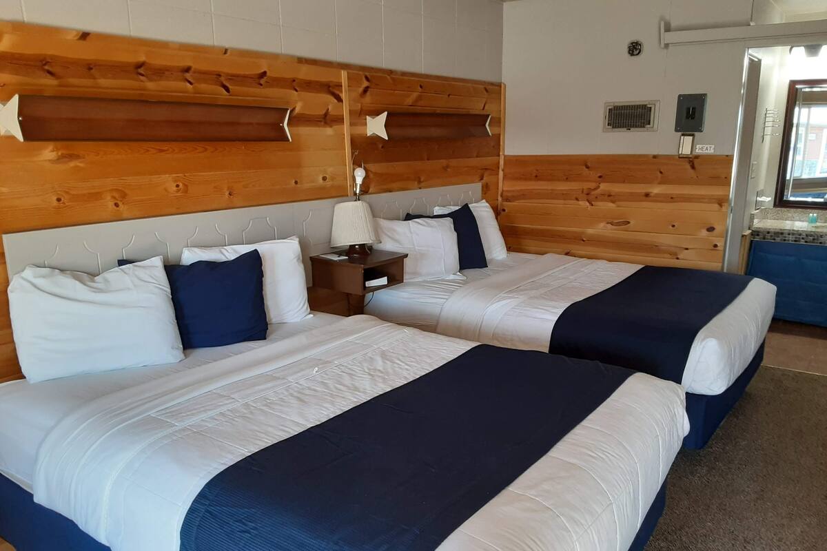 A hotel room with two beds, wooden headboard, white and navy bedding, a nightstand with a lamp, and a bathroom visible through an open door.