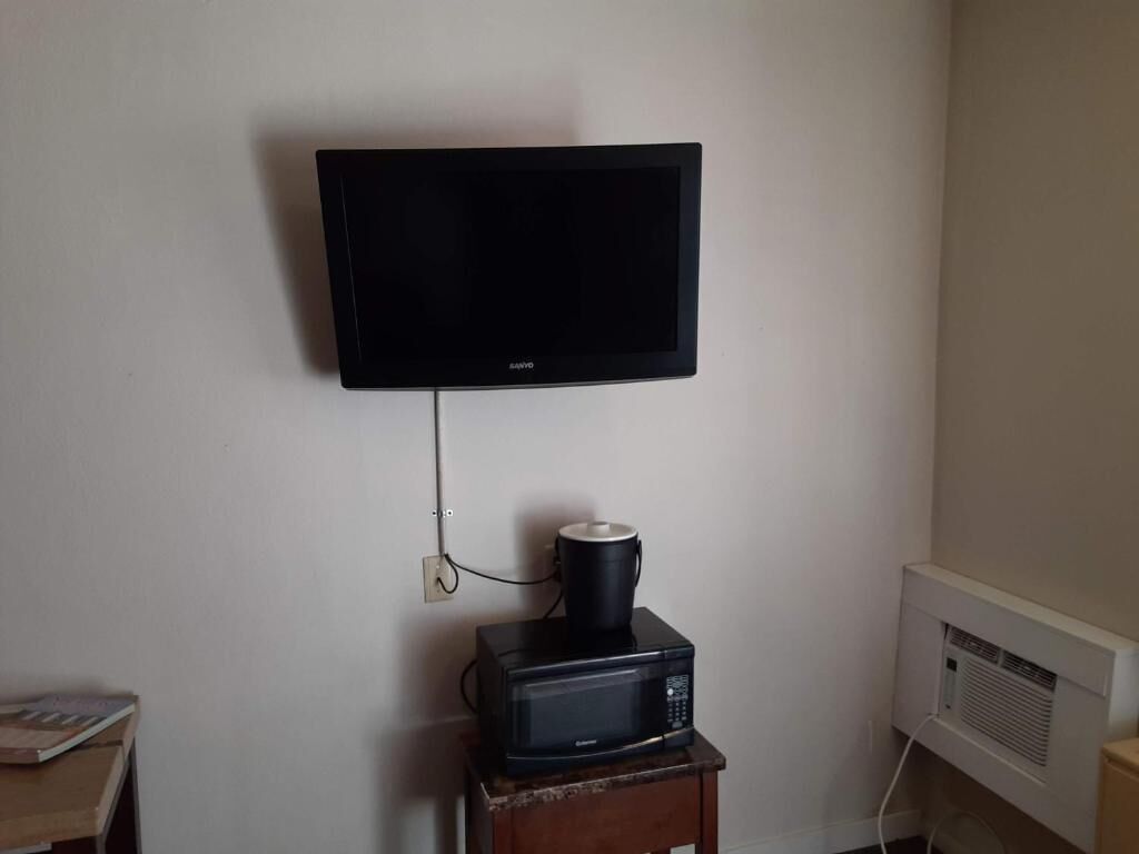 A television mounted on a wall, a microwave on a small wooden table, and an air conditioning unit on the wall.