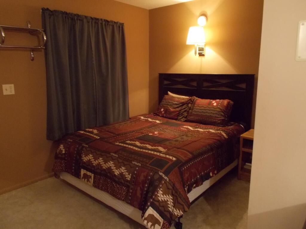 Un dormitorio con cama, mesita de noche, lámpara de pared y ventana con cortinas.