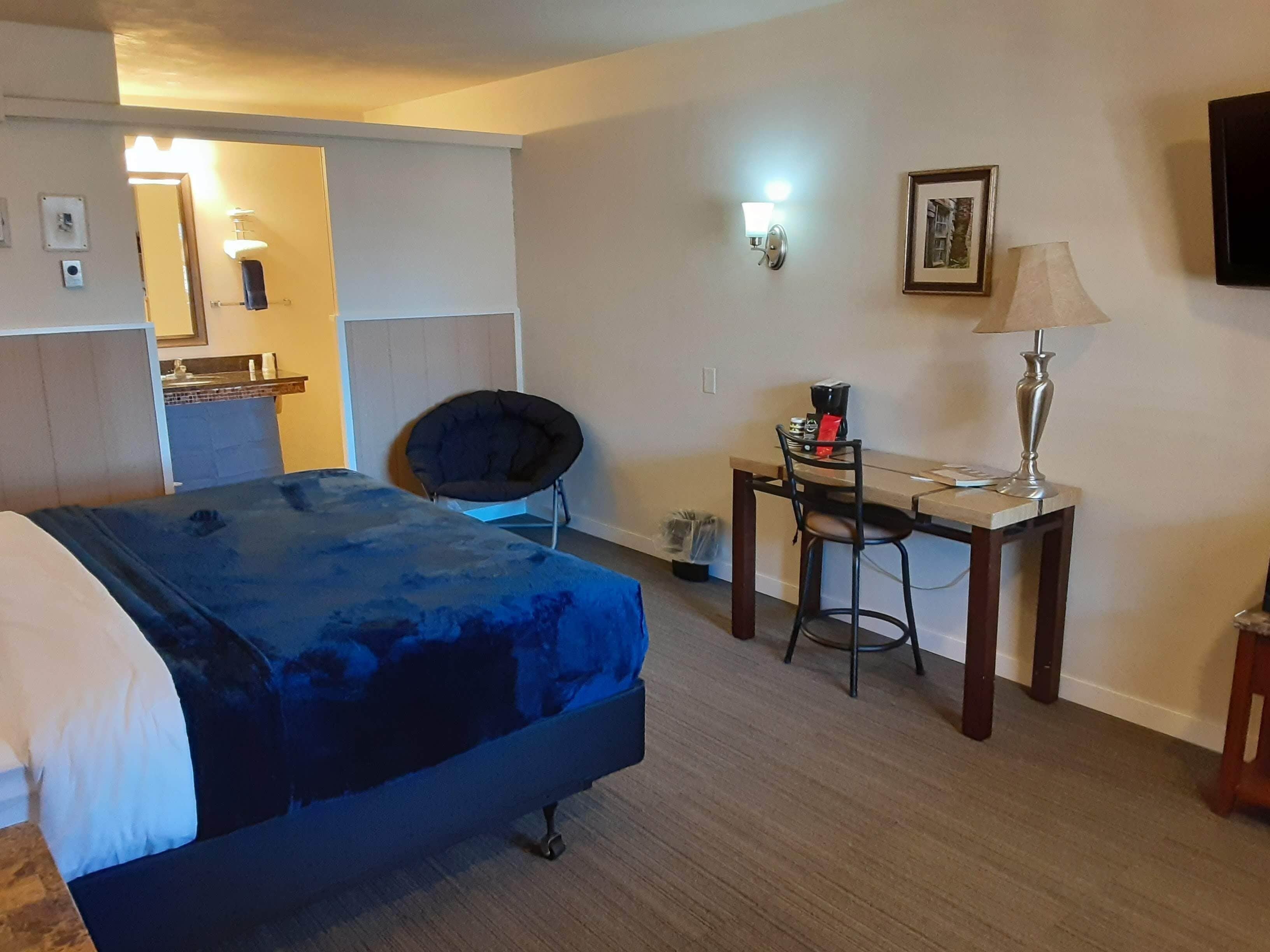 A hotel room with a bed, a desk with a chair, a lamp, and a picture on the wall.