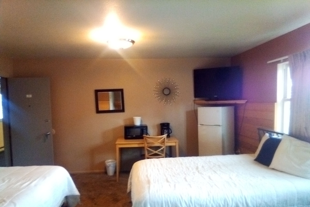 A hotel room with two beds, a desk with a computer and a coffee maker, a refrigerator, and a clock on the wall.