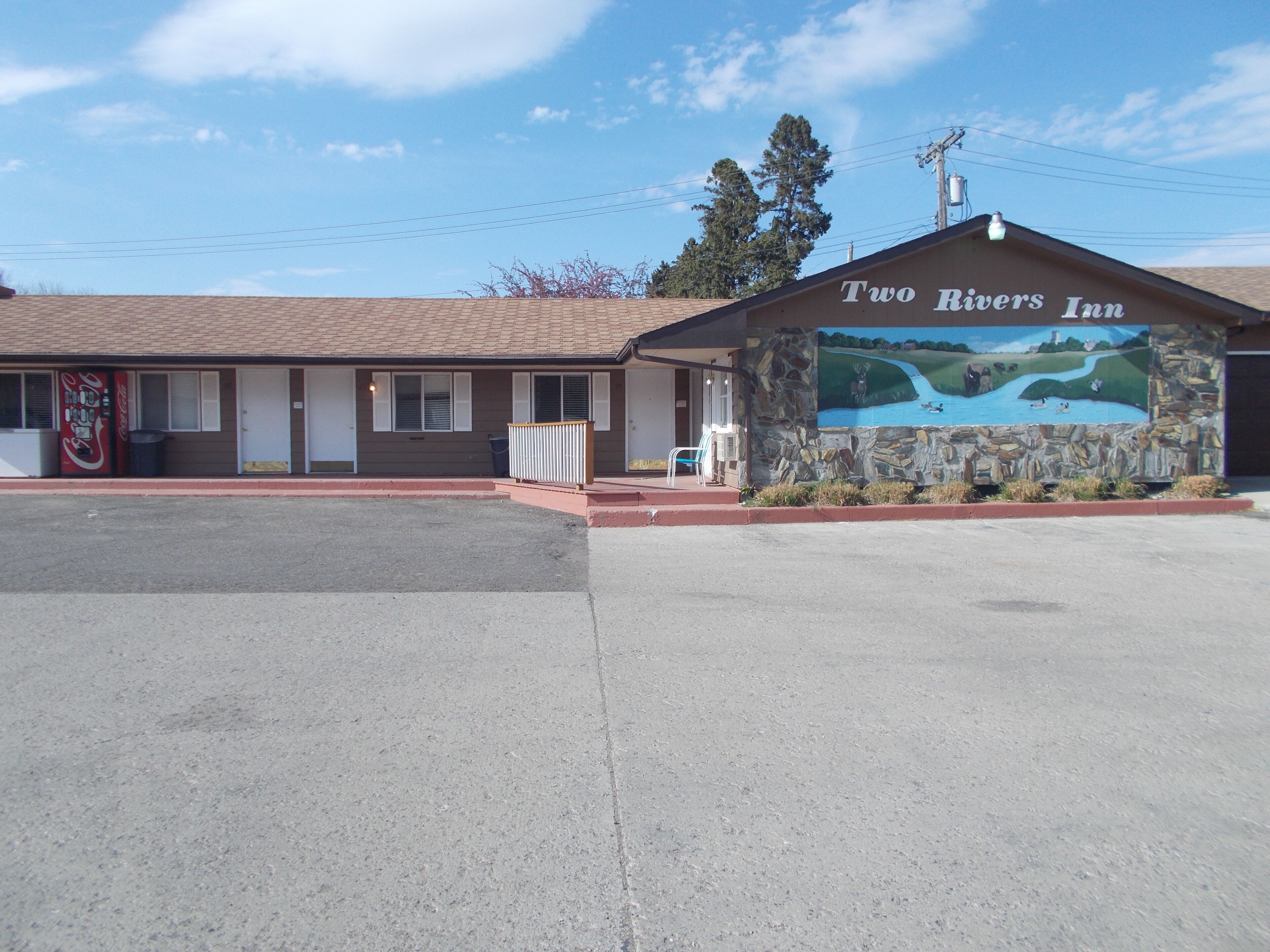 A motel named "Two Rivers Inn" with a stone facade, a Coca-Cola vending machine, and a mural of a river scene.