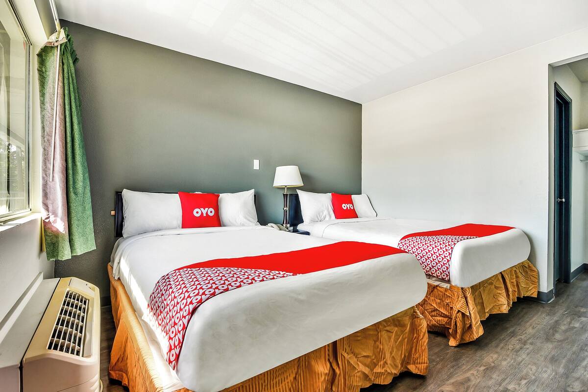 A hotel room with two beds, each with a red and white bedspread, a bedside table with a lamp, and a window with curtains.