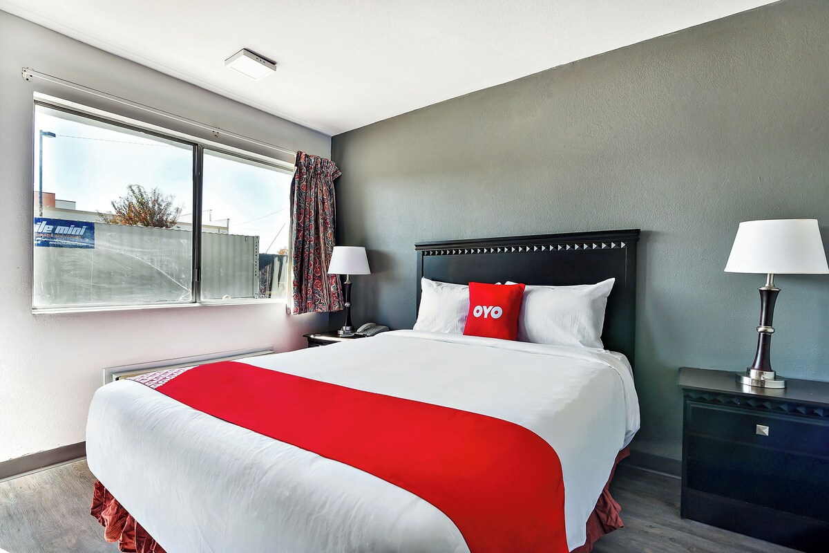 Quarto de hotel com uma cama grande, mesa de cabeceira, luminária e janela com cortinas.