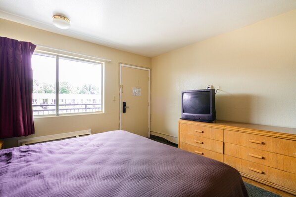Una habitación de hotel con una cama, un televisor sobre un aparador de madera, una ventana con cortinas y una puerta.