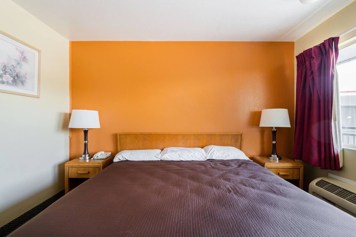 A hotel room with a large bed, two bedside tables with lamps, a nightstand with a phone, and a window with curtains.