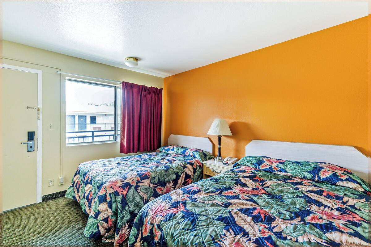 A hotel room with two beds, a window with curtains, a nightstand with a lamp, and a door.