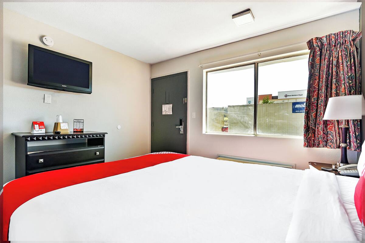 A hotel room with a bed, a TV mounted on the wall, a desk with a lamp, and a window with curtains.