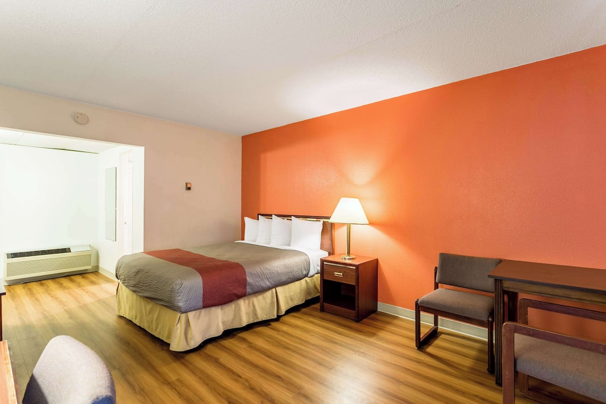 A hotel room with a bed, a nightstand with a lamp, a chair, and a wooden floor.