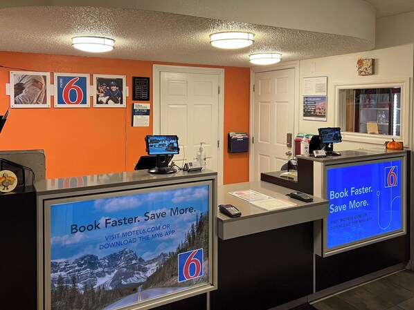 A motel reception desk with digital screens displaying promotional content, a counter with a computer, and a door to the right.