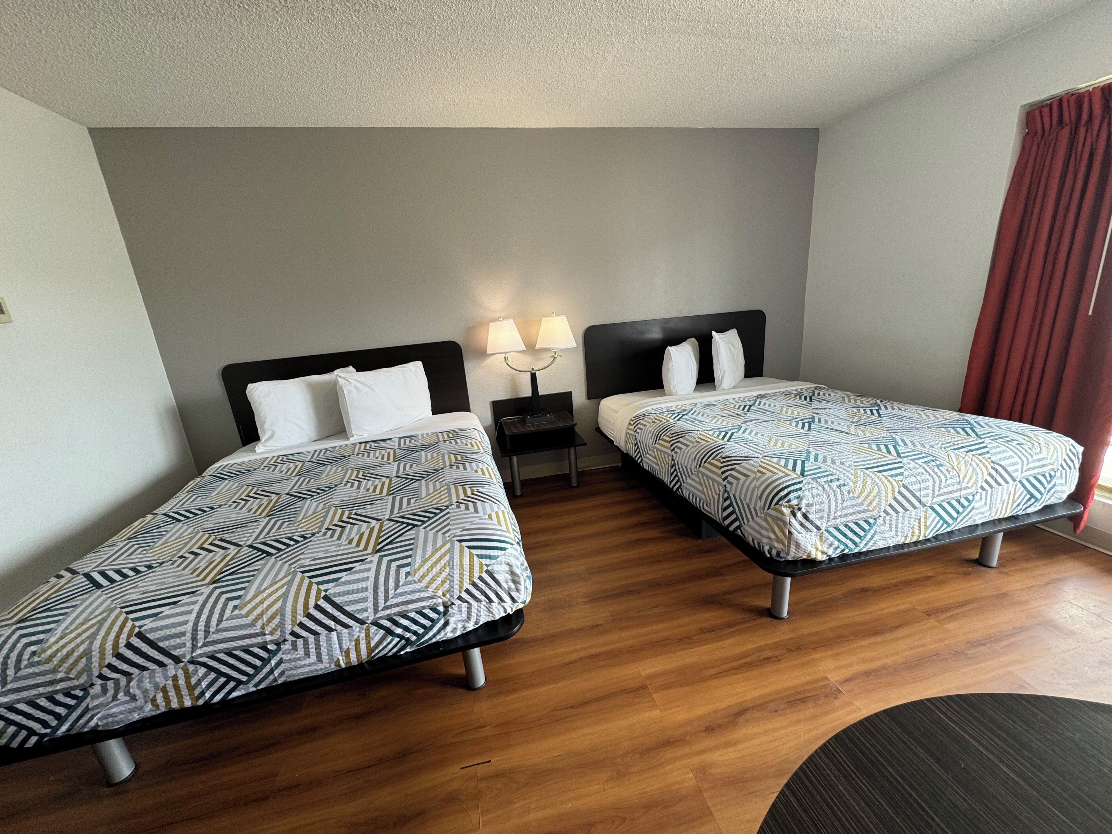 A hotel room with two single beds, a nightstand with a lamp, and a wooden floor.