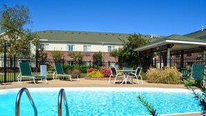A swimming pool with lounge chairs and a covered seating area, surrounded by a brick building and greenery.