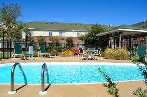 A swimming pool with lounge chairs and a covered seating area, surrounded by a brick building and greenery.